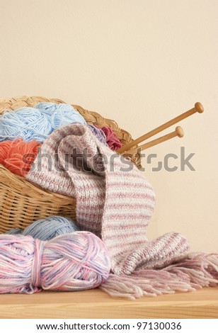 Basket with knitting yarn, wool, knitting needles and knitted scarf on a wooden table against a creamy kitchen wall. Knitting basket. Home knitting concept.