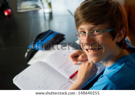 Smiling teenager studying and doing his homework while holding his pen and writing in his notebook.