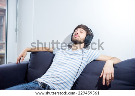 Student man listening to music on his headphones while sitting relaxed on the couch wearing a blue and white striped shirt.