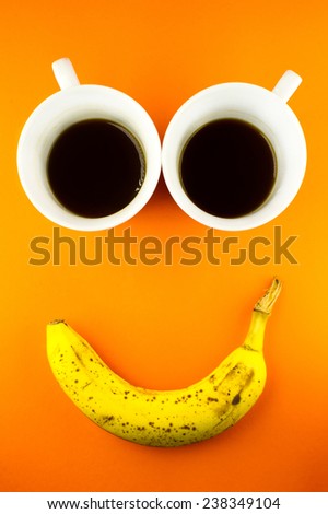 Two coffee cups and a banana shot on an bright orange background. The composition of the objects looks like a smiley face.