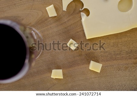 A block of cheese on a wooden cutting board shot from above. Some of the cheese is cut into small cubes. A glass of red wine is placed alongside.