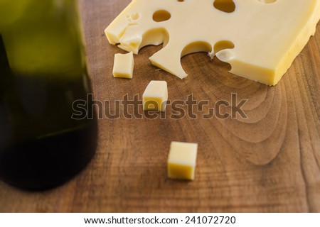 A block of cheese on a wooden cutting board shot from above. Some of the cheese is cut into small cubes. A bottle  of red wine is placed alongside.