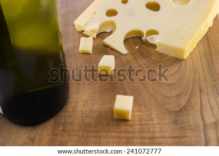 A block of cheese on a wooden cutting board shot from above. Some of the cheese is cut into small cubes. A bottle  of red wine is placed alongside.