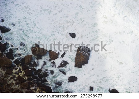 Ocean waves shot from above. The water is white and foamy. You can also see the rocky shore.