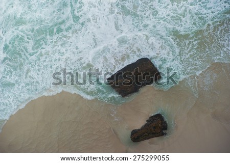Ocean waves shot from above. The water is turquoise color and you can also see a rock. You can also see the shore and sand.