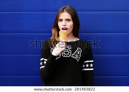 Portrait of young happy girl eating ice-cream, outdoor, over blue wall background