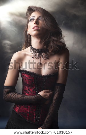 Beautiful, romantic looking girl, in a black and red lace corset with a gothic feel, long lacey gloves and a black beaded choker on a grey smokey background.