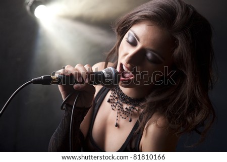 Beautiful young girl singing in a rock atmosphere wearing a black top, black lace gloves and beaded choker on a grey smokey background with a light source.
