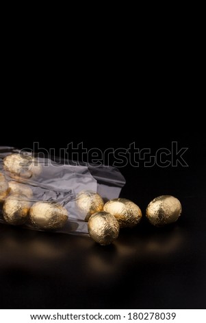 Cellophane bag of gold coloured chocolate mini easter eggs, lying down on a black surface with a couple of the eggs out of the bag. Focus is on the front egg.