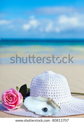 On sand before the sea the rose flower, a  hat and black pearls of Tahiti in a shell lies.