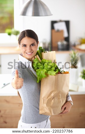 Young woman holding grocery shopping bag with vegetables and showing ok
