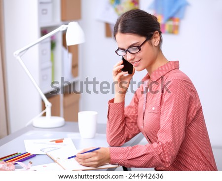 Young attractive female fashion designer working at office desk, drawing while talking on mobile