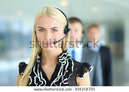 Businesswoman with headset smiling at camera in call center. Businessmen in headsets on background