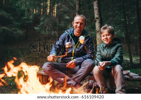 Father and son roast marshmallow candies on the campfire in forest. Spring or autumn camping