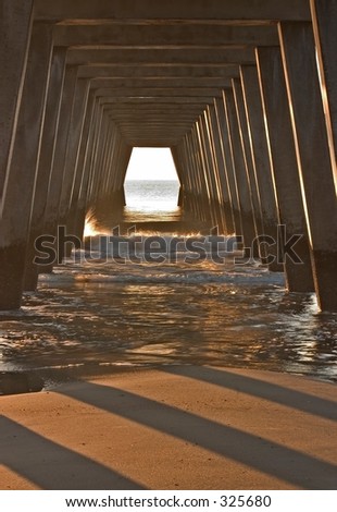 Looking out toward the ocean in early morning light under a pier on a beach at low tide.