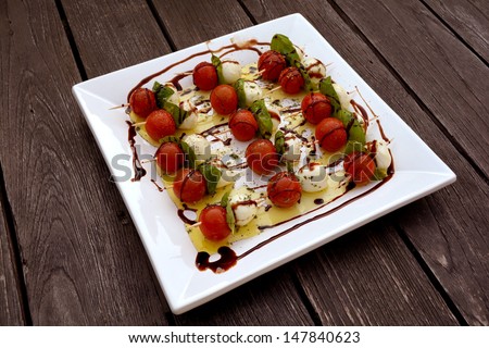 A square white plate with sticks of cherry tomatoes, Italian Mozarella cheese balls and fresh basil leaves topped with olive oil and balsamic vinegar served as starter on an old wooden table outside.