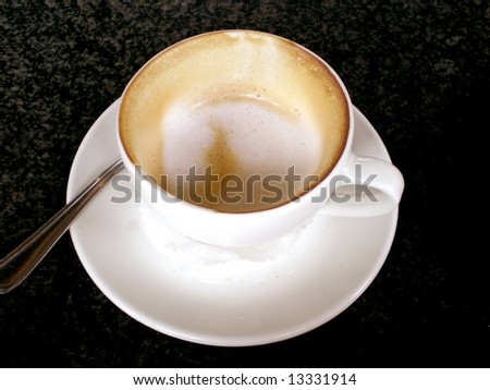 A white cup of an almost finished delicious Italian skinny (low fat) Cappuccino with silver spoon standing on a black granite table of a coffee-shop outdoors