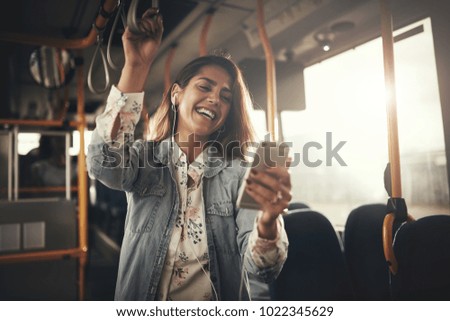 Young woman wearing earphones laughing at a text message on her cellphone while riding on a bus