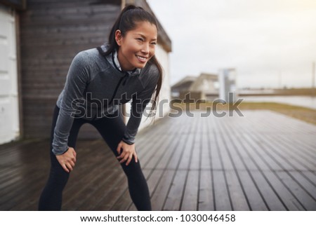 Smiling young Asian woman in exercise clothing standing with her hands on her knees while taking a break from a run on an overcast day