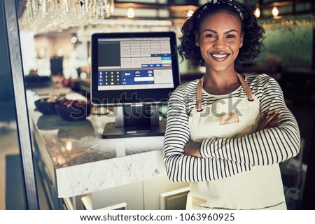 Young African waitress wearing an apron and smiling while standing by a point of sale terminal in a restaurant