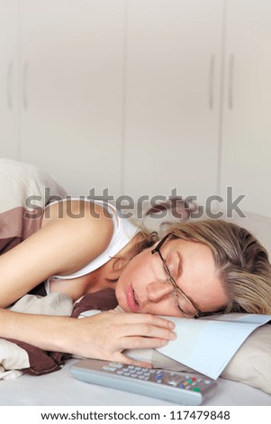 Exhausted woman sleeping in her glasses slumped over the paper that she had been reading while lying in her bed