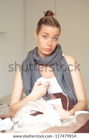 Beautiful young woman with flu feeling sorry for herself sitting in bed with a scarf around her neck and a toilet roll for tissues