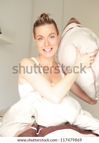 Smiling beautiful young woman sitting in bed taking aim with a pillow during a mock pillow fight