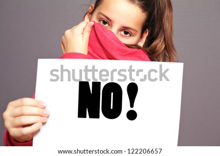 Unhappy little girl hiding behind the collar of her jacket saying an emphatic NO by means of bold type text on a sheet of white paper