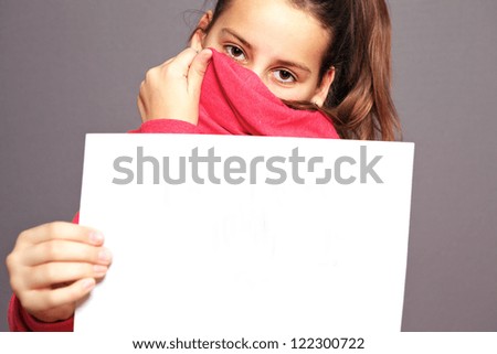 Bashful little girl hiding her face with the collar of her jacket so that just her eyes are visible holding up a blank sheet of white paper