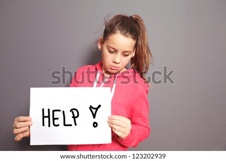 Conceptual image of a sad dejected little girl with a pouting lip standing holding a handwritten HELP sign with an exclamation mark, studio upper body portrait on a grey background with copyspace