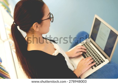 High angle over the shoulder view of a young woman wearing glasses sitting typing on her laptop balanced on her lap