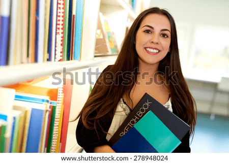 Happy young art student at the college library standing alongside a bookcase full of books with a sketchbook clasped in her arms smiling at the camera