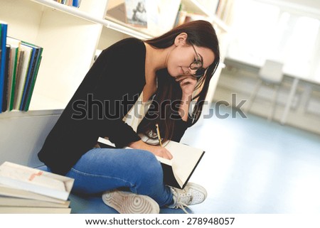 Young woman sitting on the floor against a bookcase full of books working in a college library taking notes as she researches a class project