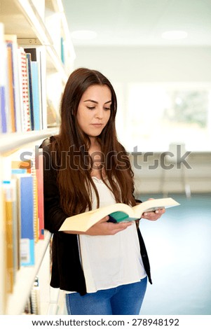 Pretty young female university student doing research in the library standing leaning against a bookshelf reading a book