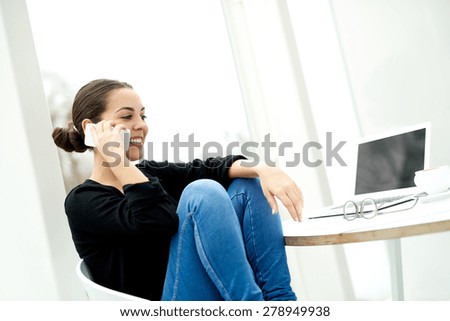 Pretty young woman relaxing with her mobile phone sitting with her feet up on her chair smiling as she chats with a friend