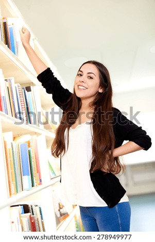 Young woman reaching for a book from a tall wall mounted book case smiling as she looks at the camera