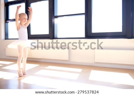 Little girl training to be a ballerina standing in a graceful arms raised position in her white tutu and pink satin ballet shoes in a bright sunlit ballet studio