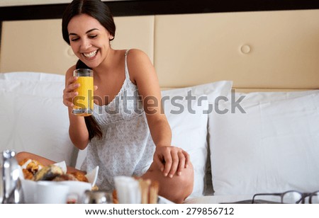 Attractive woman wearing nightgown relaxing in bed holding glass of orange juice from breakfast tray.