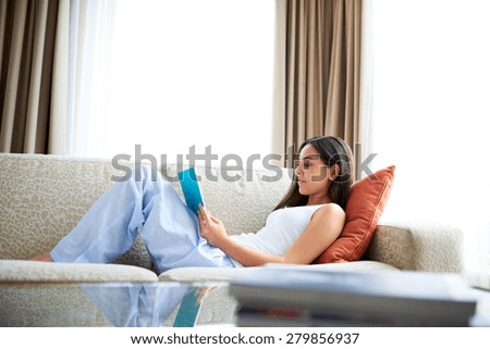 Woman reclining on orange cushion on arm of couch reading a book.