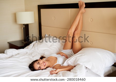 Attractive woman smiling lying upside down on bed with legs resting on headboard.