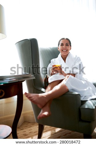 Young woman sitting in chair with legs over chair arm holding glass of orange juice.