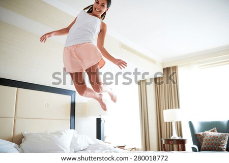 Angled view of woman jumping on bed wearing pajamas and smiling at camera.