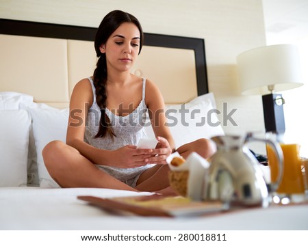 Attractive woman relaxing in bed with breakfast tray and looking down at cell phone.