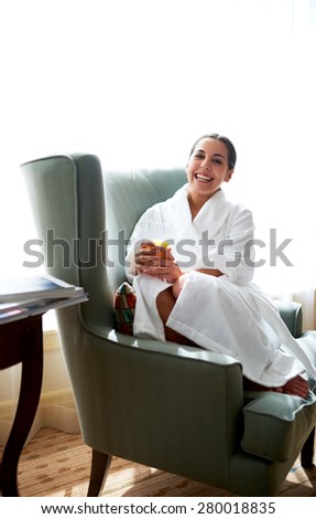 Young woman curled up in chair holding a glass of ornage juice and smiling.