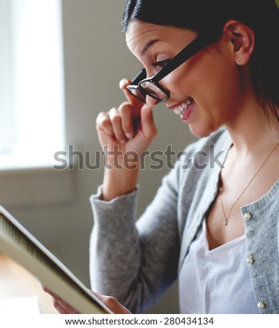 Woman smiling and pulling glasses down nose while reading book in home office.