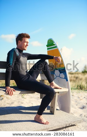 Attractive male surfer wearing his wetsuit sitting relaxing on a wooden bench with his surfboard on top of a sand-dune and a sandy beach on a hot summer day