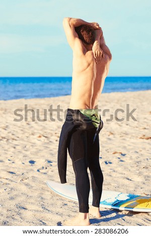 Surfer reaching for his zipper string pulling it up over his shoulder to zip up his wetsuit as he stands on a tropical beach with his surfboard facing the ocean