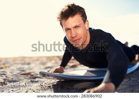 Close up Handsome Young Male Surfer Lying on his Stomach on Top his Surfboard at the Beach Sand, While Smiling at the Camera.