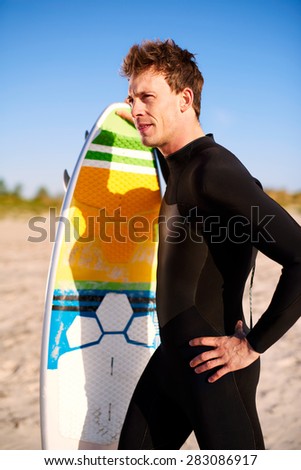 Athletic fit young surfer with his surfboard standing on a tropical beach holding his board upright staring out over the ocean, close up side view in summer sunshine