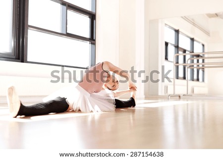 Young Pretty Ballet Dancer Doing Stretching Exercise on the Floor Inside the Studio with Legs Split and Body Bent on her Side.